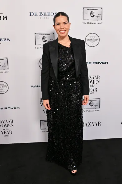 Guest wearing a black sequin floor-length gown with a tailored black blazer, posing on the Harper’s Bazaar Women of the Year 2025 red carpet in front of a step-and-repeat backdrop.