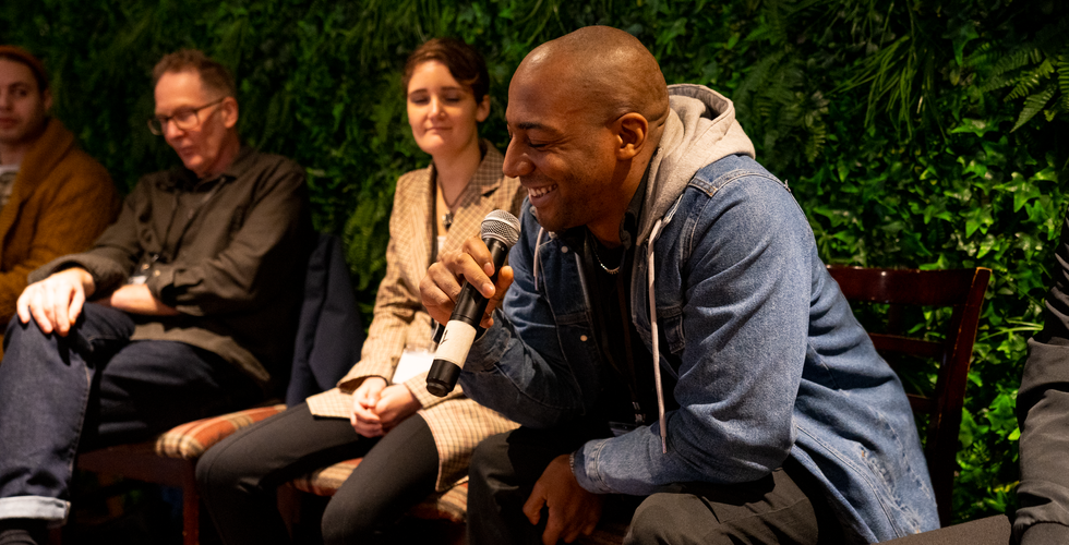 “Participant speaking into a handheld microphone during a panel discussion, seated in front of a green plant wall.”