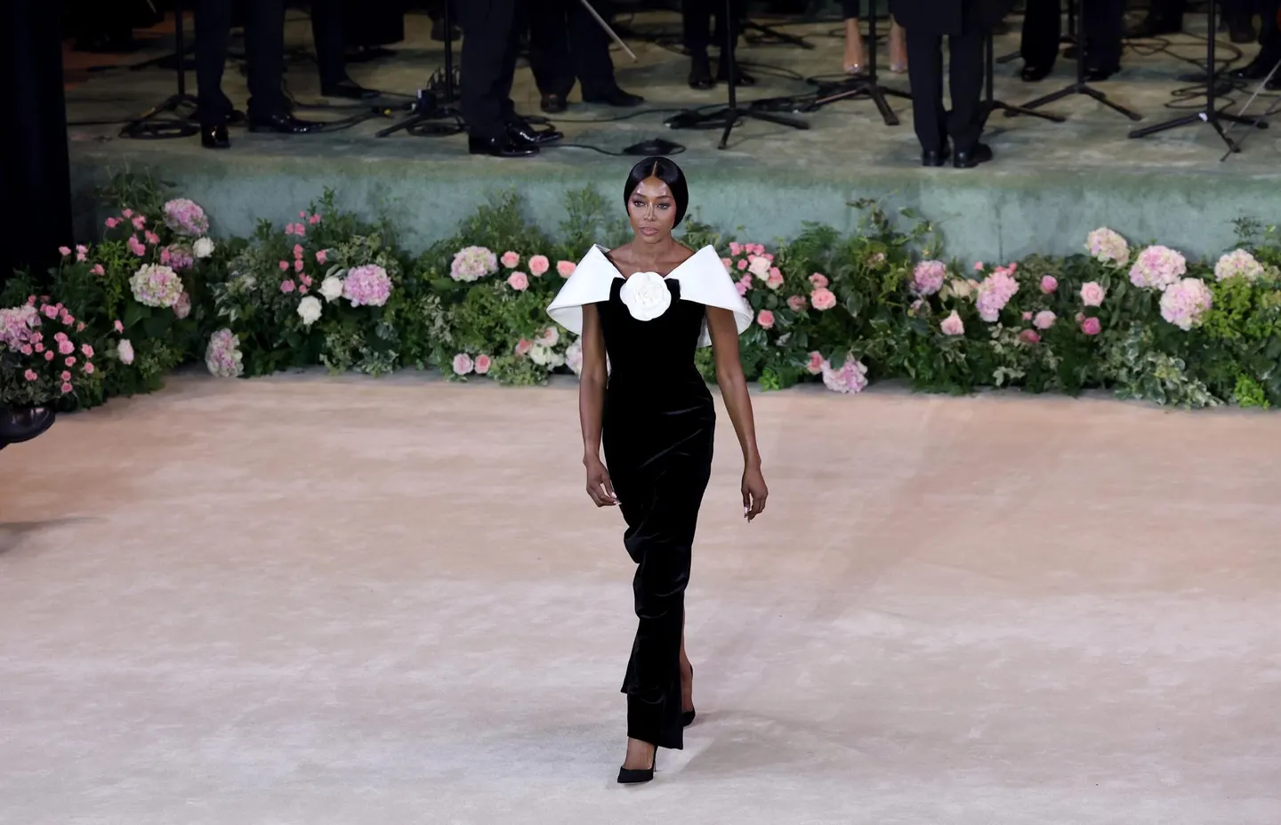 A model in a black velvet gown with bold white shoulder detail and a large flower motif walking a floral-lined runway during an elegant fashion presentation.