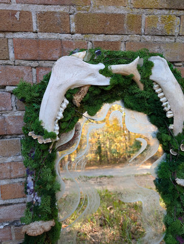 A close up of the top half of the Forest Floor Portal Mirror, with the elk jawbones and deer antler creating an arch. The golden glow of the woods can be seen in the reflection of the mirror.