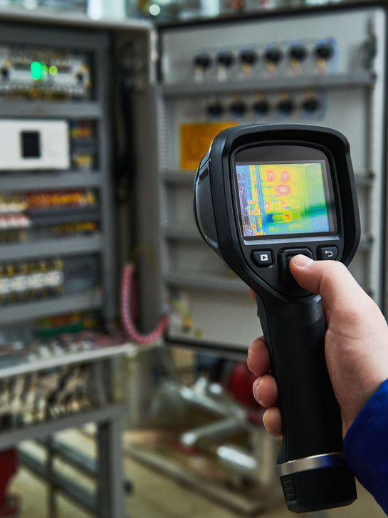 A thermal camera held up to an open electrical control cabinet.