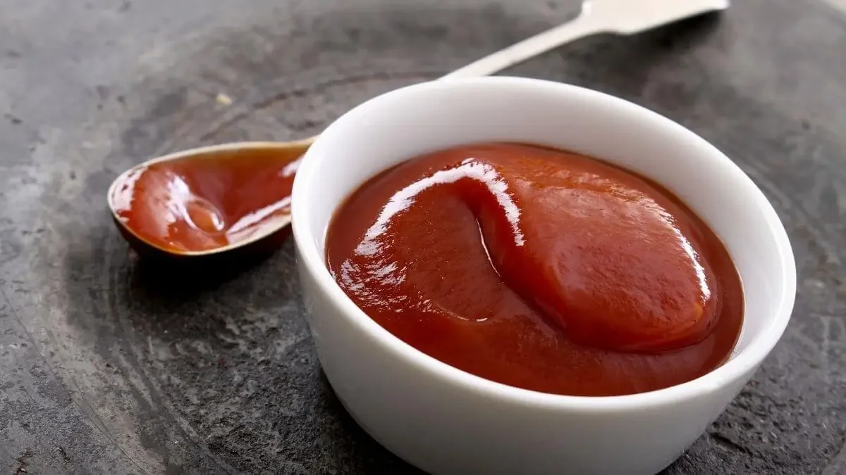 Um potinho de porcelana branca com katchup caseiro sobre uma mesa rústica de pedra escura, ao lado esquerdo, uma colher suja com o molho de ketchup