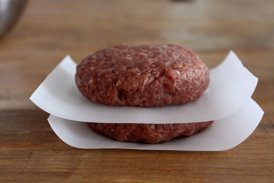 Foto de dois hamburgueres bovinos caseiros, um em cima do outro em uma mesa, separados com uma folha de papel manteiga