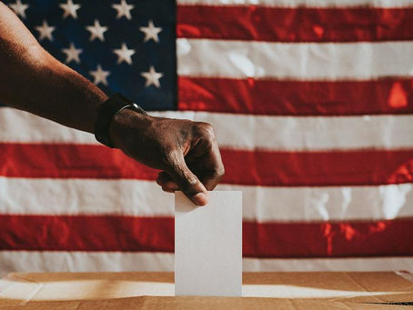 A Black man in the foreground, dropping a paper ballot into a ballot box, with a large American flag being featured in the background.
