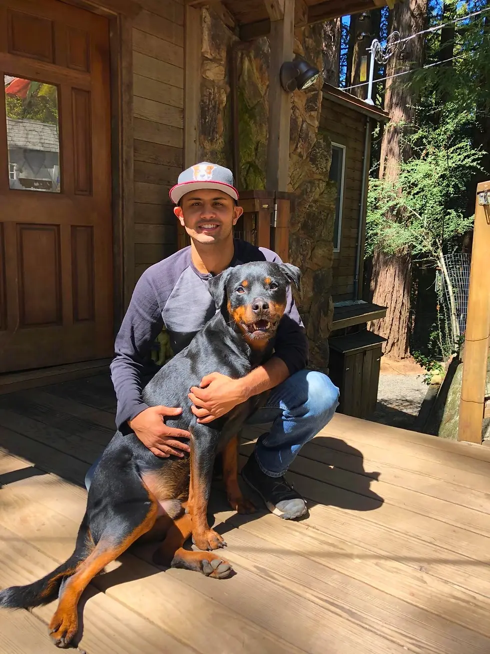 Young man wearing a gray baseball cap and black long-sleeve shirt kneels on a wooden porch, smiling and hugging a large Rottweiler dog. They are in front of a rustic cabin surrounded by trees on a sunny day. The scene conveys warmth and companionship in an outdoor, nature setting.