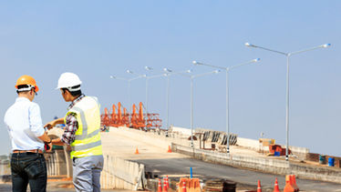 Two construction workers in hard hats reviewing plans at a road or bridge construction site with barriers, cones, and equipment under a clear blue sky.