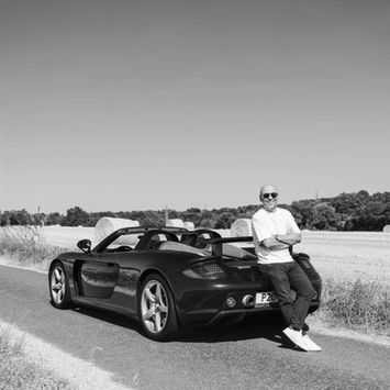 Man leaning against a black Porsche on a country road.