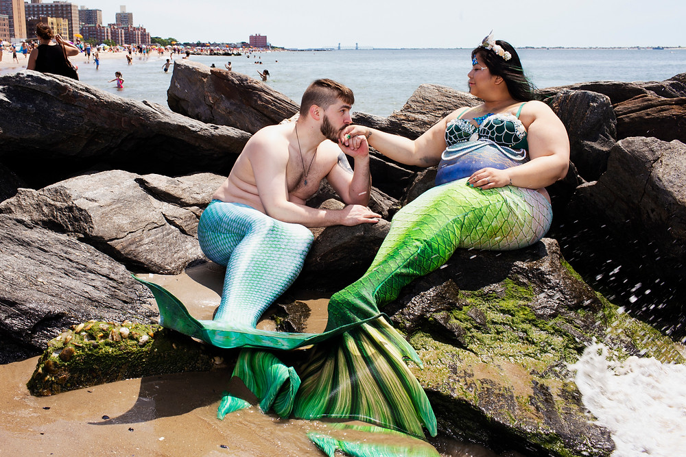 two merpeople sitting on a rock. One is pregnant. The merman is kissing the mermaid's hand