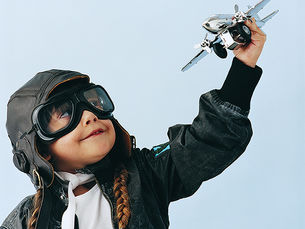 Child wearing aviator goggles and a cap, holding a toy airplane up, smiling. Dressed in a leather jacket with braids. Blue background.