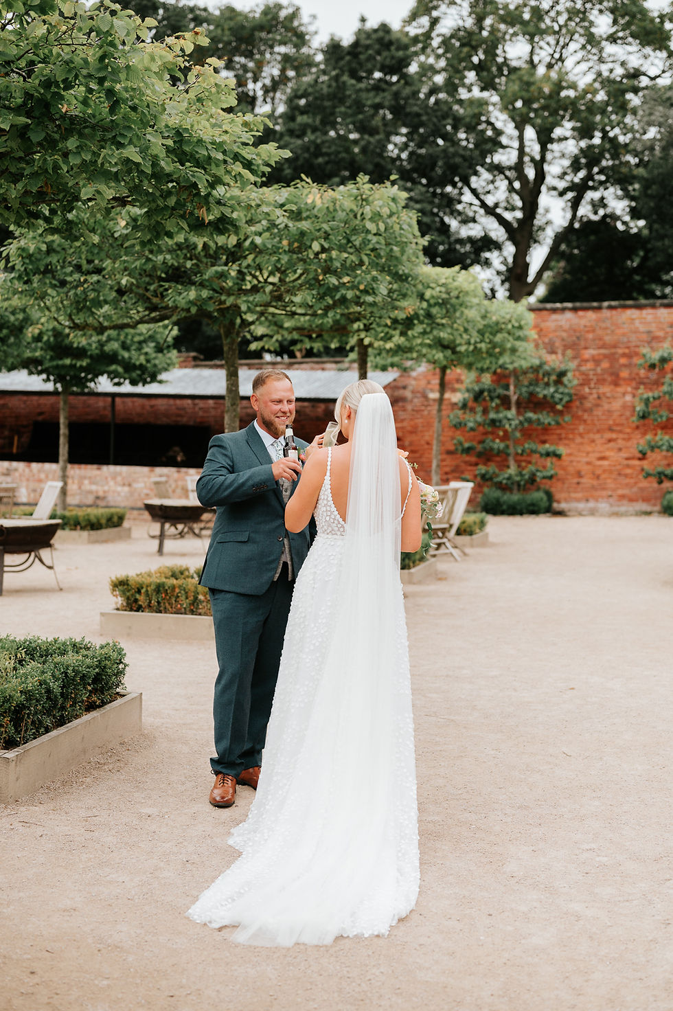 Bride in white dress and groom in a suit toast with drinks in a garden setting with trees and brick walls, conveying joy and celebration.