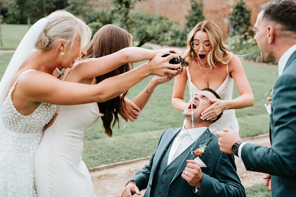A playful outdoor scene shows a bride and friends pouring champagne on a laughing groom's face, surrounded by greenery and joy.