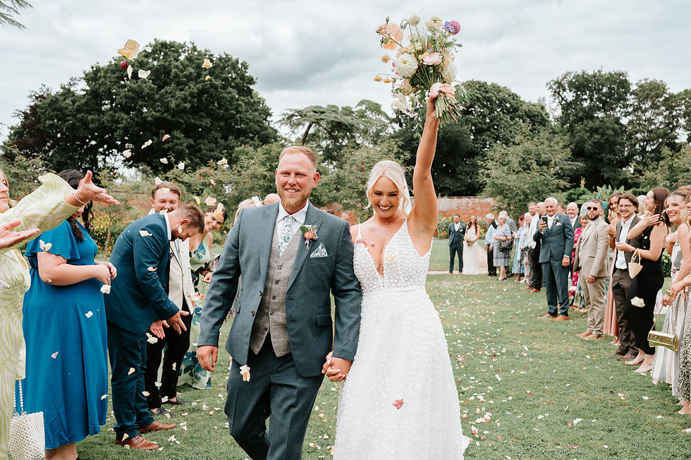 Bride and groom walk hand in hand outdoors, smiling. Guests cheer and throw petals. Bride holds bouquet aloft. Lush greenery surrounds.