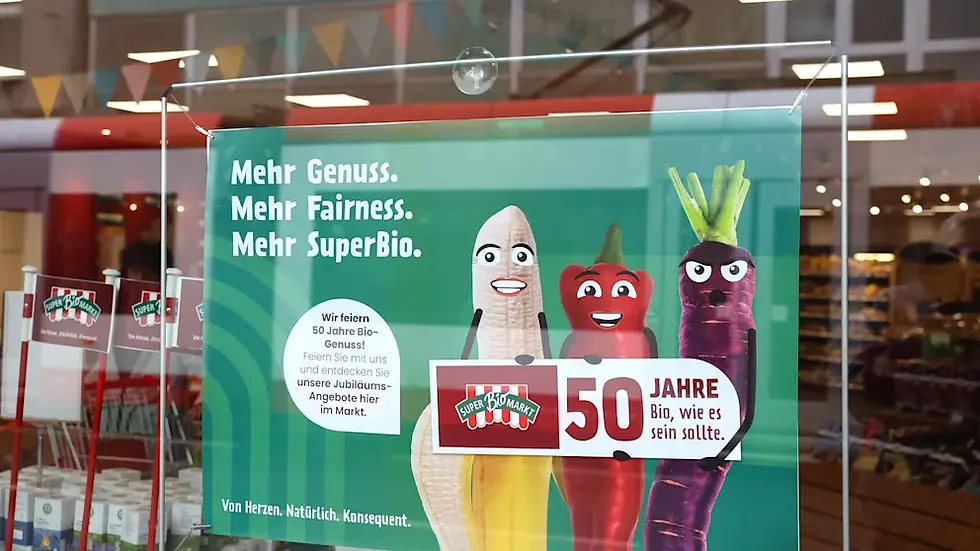 Ein Werbeplakat zeigt drei lustige, anthropomorphisierte Gemüse (eine Karotte, ein Rote Bete und ein Lauch), die vor einem grünen Hintergrund mit dem Text „Mehr Genuss. Mehr Fairness. Mehr SuperBio.“ stehen. Das Plakat feiert das 50-jährige Jubiläum der Bio-Produkte mit dem Slogan „50 Jahre Bio, wie es sein sollte“ und hebt die nachhaltigen, fairen Angebote hervor.