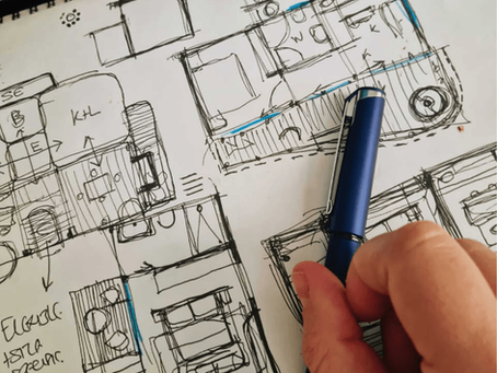 Hand holding a blue pen over a detailed sketch of architectural floor plans, featuring rooms with labeled sections and highlighted areas.