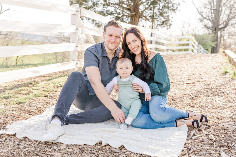 Spring Family Mini Portrait Session of a family with a baby boy at Ellington Agricultural Center in Nashville, Tennessee with Family and Newborn Photographer Janet Henry Photography. Baby is wearing soft muted green overalls with a long sleeve white shirt. Mom is wearing jeans and a blue green blouse. Dad is wearing a gray polo with gray pants and white shoes
