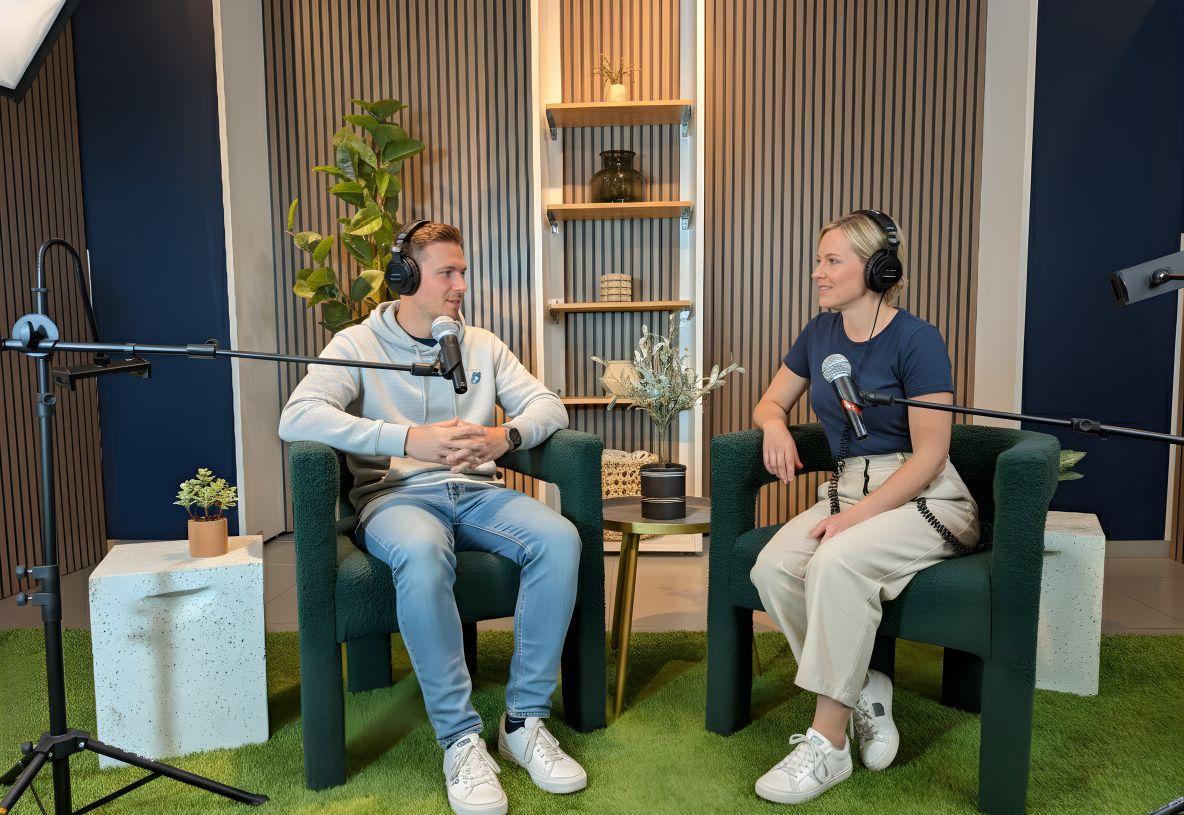 Two people with headphones sit and talk on a podcast interview set.