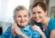 elderly diabled woman in chair smiling while a yonger Direct Support Professional is slightly hugging her smiling. The elderly woman holds the hand of the younger woman with comfort.