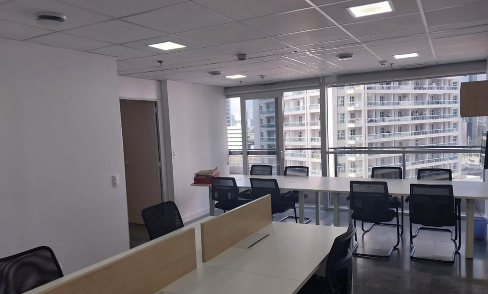 "Interior de uma sala mobiliada com mesas e cadeiras de escritório modernas. A sala é bem iluminada, com luzes embutidas no teto e grandes janelas que oferecem vista para outros edifícios próximos. As paredes são brancas e há uma porta fechada ao fundo à esquerda."