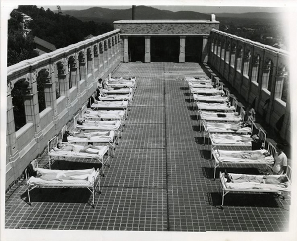 Black-and-white rooftop terrace with rows of people lying on cots, like a sanatorium, under open sky and distant hills.