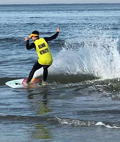 Kai Owens riding his skimboard wearing a yellow vest over his wetsuit that has the words blind athlete on it.