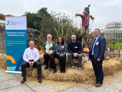 A group of people sitting on hay bales in front of a scarecrow at the National Botanic Gardens in Glasnevin. The greenhouse is visible in the background and there is a pull-up banner which reads "Central Statistics Office - Ecosystem Accounts Stakeholder Forum - Thursday 16 October 2025, National Botanic Gardens Dublin".