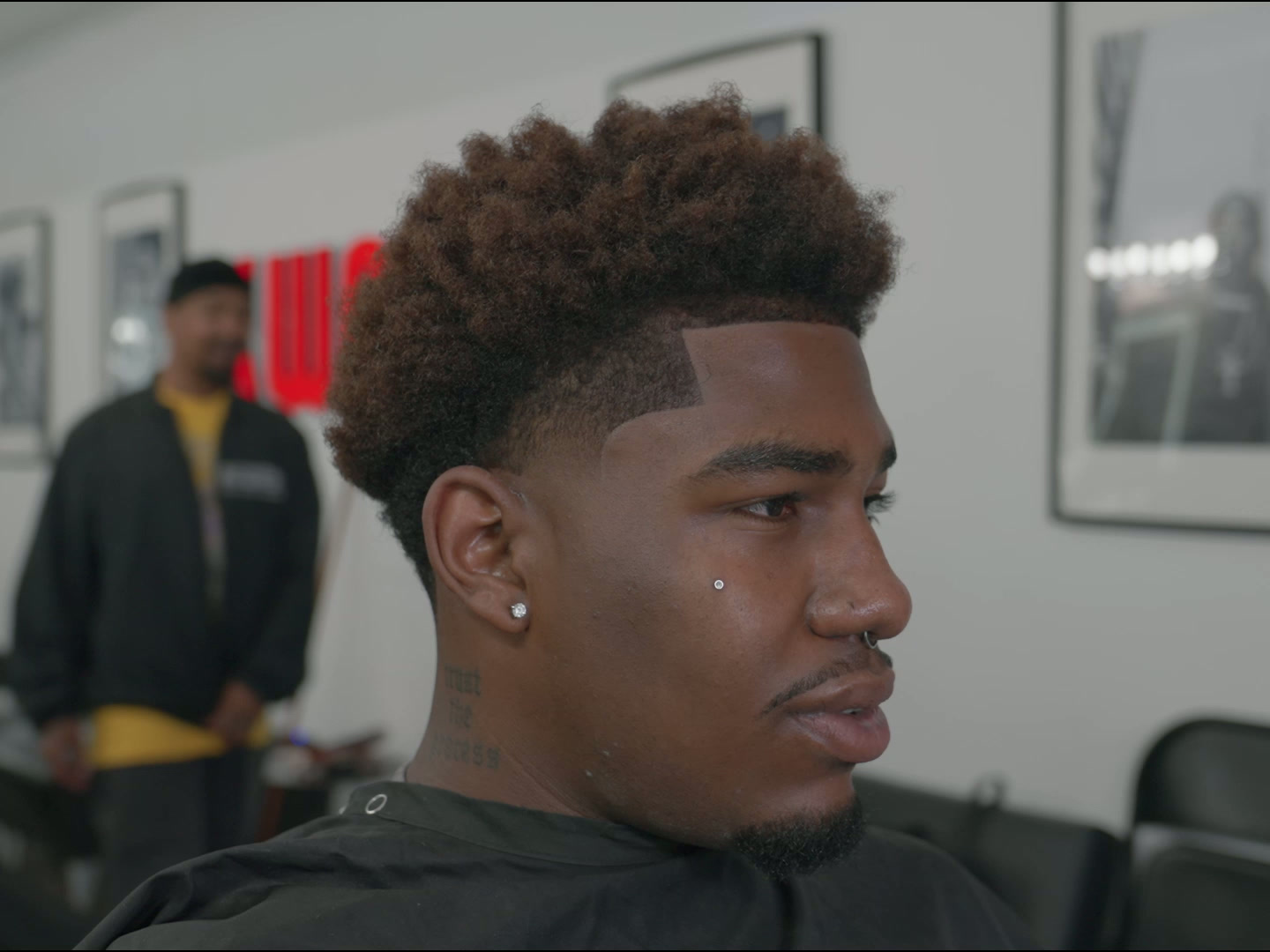 Curly top with sharp part and fade, barber visible in background