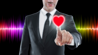 A man in a business suit stands in front of a soundwave in all the colors of the visible spectrum. A red heart appears on the tip of his left index finger as he points it up towards the viewer.
