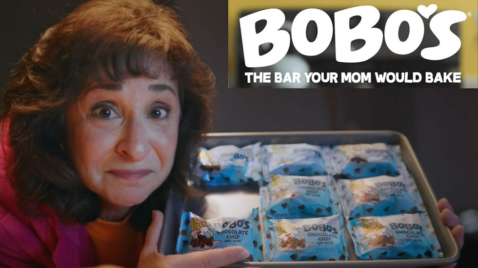 Actor Laura Doman with a tray of Bobo's chocolate chip oat bites in blue packaging, smiling gently. Text: Bobo's, The Bar Your Mom Would Bake.