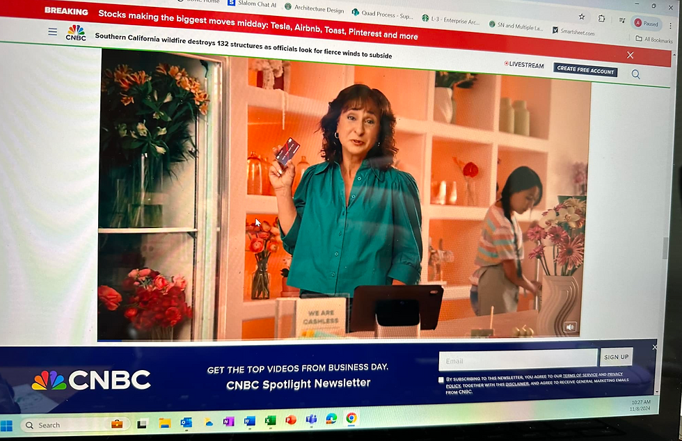 Actor Laura Doman (woman in teal) holds a card in a flower shop, with a sign "WE ARE CASHLESS." A young girl behind her arranges flowers. Bright, colorful setting.