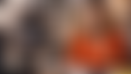 Woman in orange shirt speaks energetically during a video recording. Microphone and camera visible. Cozy room with warm lighting in background.