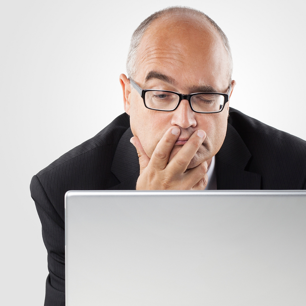 Bald man in glasses wearing a suit looks focused at a laptop. He has a thoughtful expression, hand on chin. Plain white background.