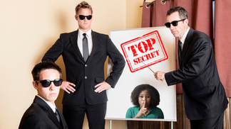 Three secret service men stare back at the viewer while one uses a pointer at flip chart that says Top Secret and shows a woman with her finger against her lips, indicating quiet.