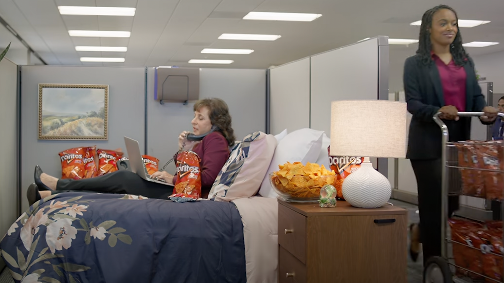 Actor Laura Doman working in bed in her office cubicle eats Doritos, using laptop and phone. Another woman pushes a cart of chips. Lamp, framed art, and floral bedding.