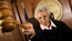 A judge in her robes and courtroom extends her gavel towards the viewer with a stern expression.
