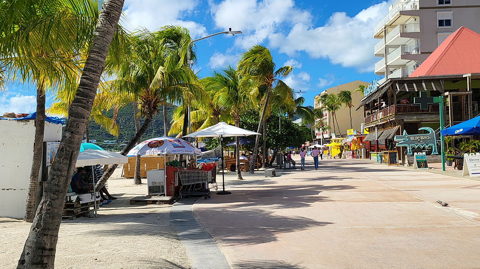 Beachfront street lined with palm trees; people walking, vibrant storefronts and colorful umbrellas under a clear blue sky. Relaxed atmosphere.
