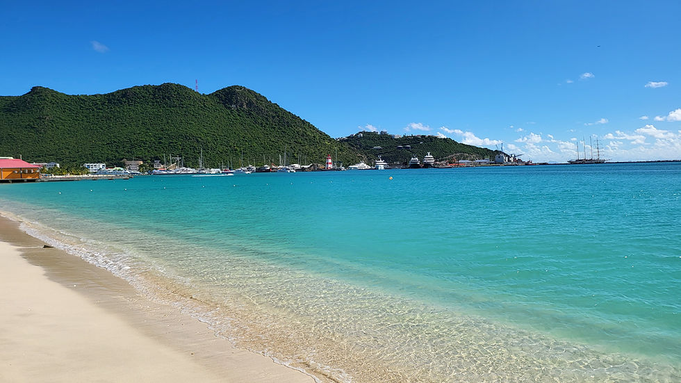 Beach view with turquoise water, sandy shore, and distant green hills. Boats and a red lighthouse are visible under a clear blue sky.