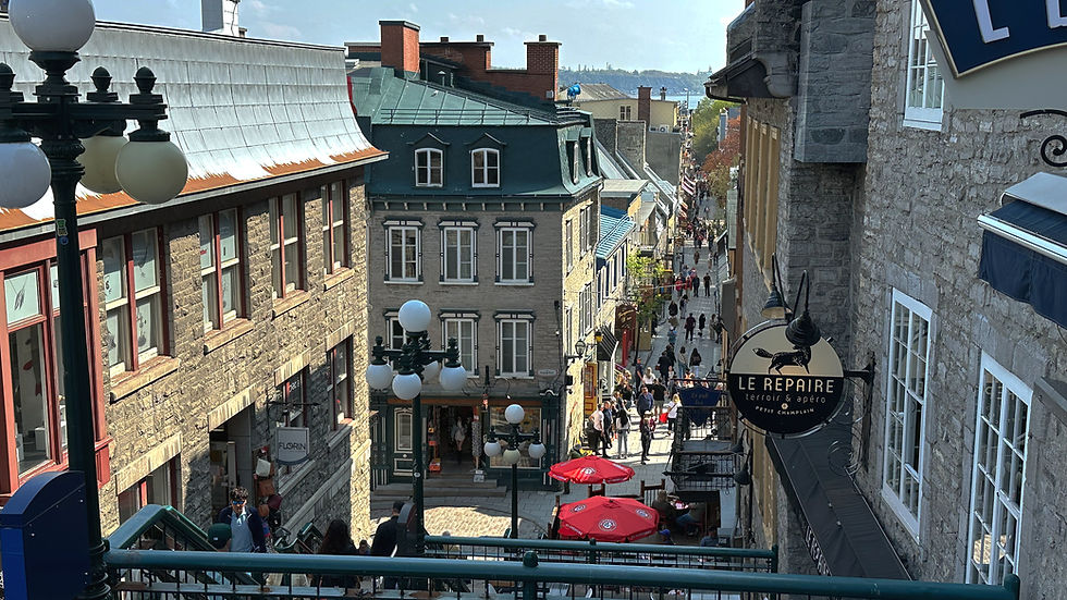 Narrow street in Quebec City with people walking, bordered by stone buildings. Red umbrellas and "Le Repaire" sign visible. Bright, sunny atmosphere.