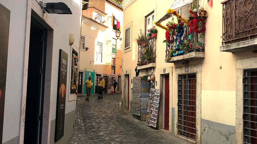 Narrow cobblestone street with colorful buildings, flower-adorned balconies, and people walking. Warm light creates a cozy, inviting atmosphere.