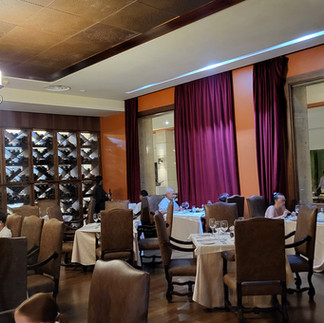 a restaurant with burgundy curtains, a few people seated at tables with brown chairs; bottles arranged on shelves in the background