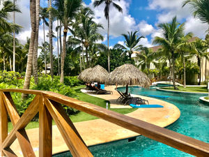 Tropical resort pool with palm trees, thatched umbrellas, and lounge chairs. Wooden bridge in foreground. Bright, sunny day mood.