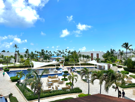 esort with palm trees and white buildings, featuring pools, shaded seating areas, and people walking. Clear blue sky and tropical vibe.