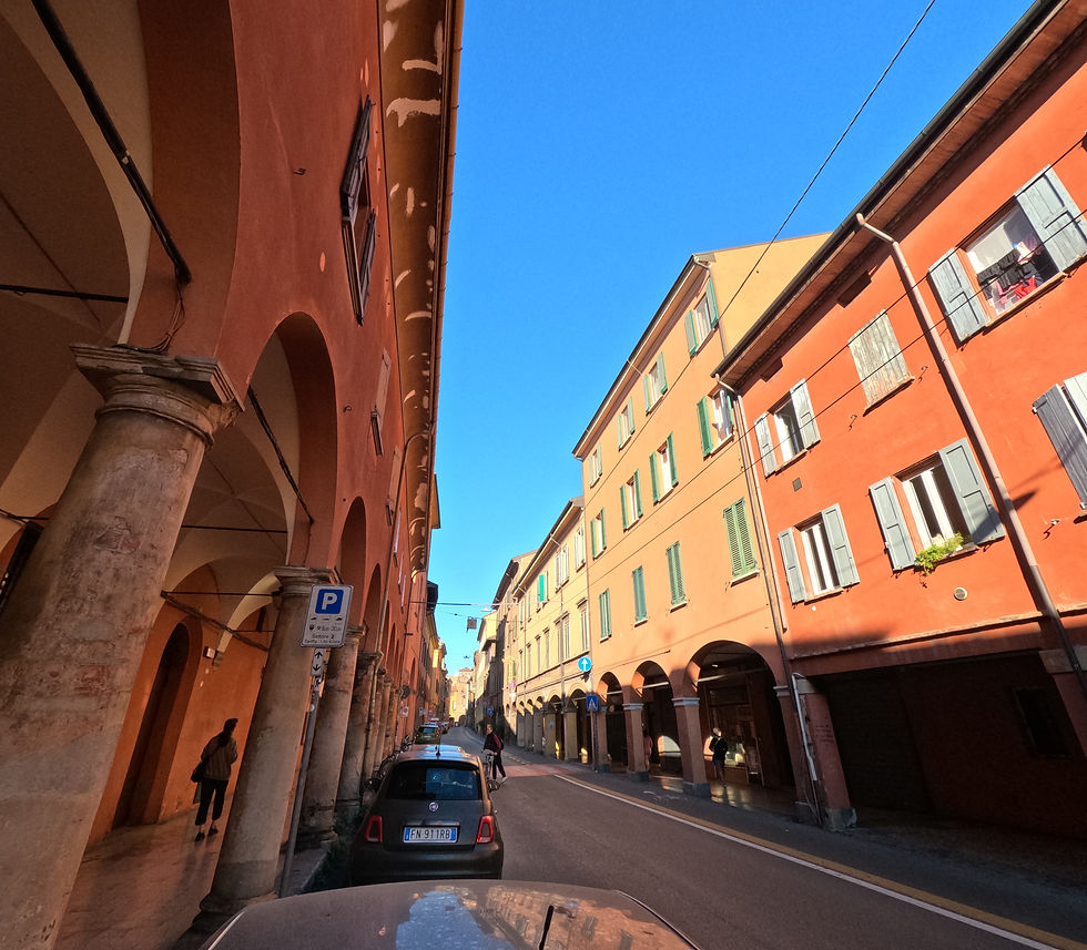 Narrow street in Bologna with red-orange buildings and arched walkways (porticoes) under a blue sky. People walk on sidewalks, and cars are parked along the road.