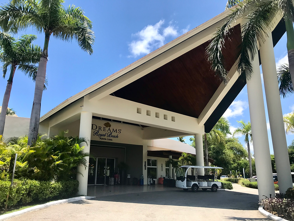Entrance of Dreams Royal Beach, Punta Cana. A white golf cart is parked near tall columns. Lush greenery and palm trees under a clear blue sky.