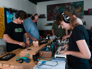 Teilnehmende des Workshops „Rhythmus & Synthese“ sitzen konzentriert an einem Tisch mit verschiedenen Synthesizern und Drummachines. Jede:r Teilnehmende justiert einen Regler, während sie den Veränderungen der eigene Klänge lauschen und neue Sounds ausprobieren.