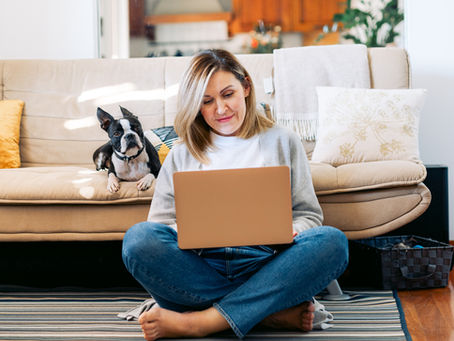 Woman working casually on a laptop while seated on the floor, with her dog nearby on the couch.