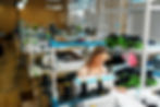 Female garment worker sewing clothes in a modern textile production workshop with stacks of apparel ready for bulk clothing production.