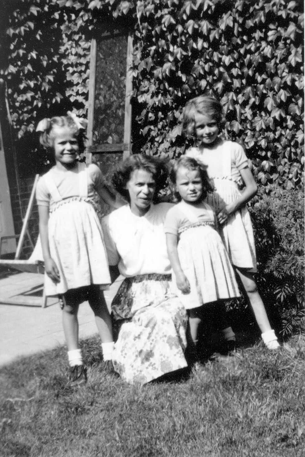 A black and white photo of my mother, my two aunts, and my oma. It was taken outdoors, and all four of them are squinting into the sunlight. The girls are all wearing the same dresses made by my oma. She is kneeling in grass with her arms around the younger two girls, while all three girls are standing. They range from about three to eight years old. They are outdoors and posing for the camera. The photo was taken in about 1950.