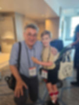 An adult man kneels beside a smiling child wearing leg braces. Both wear conference badges with lanyards. They are indoors at the conference, with other attendees and a stroller in the background.
