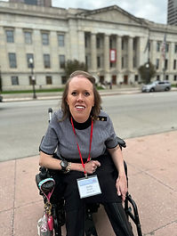 Kelly sitting in her wheelchair, smiling with light brown hair, wearing a gray and black sweater with black pants, in front of the Ohio State Capitol Building located in Columbus, Ohio.