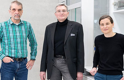 Three adults standing in a laboratory setting. One person on the left wears a green plaid shirt and jeans, the person in the center wears a black turtleneck with a dark blazer, and the person on the right wears glasses and a black sweater while leaning slightly against a lab bench. Shelving, cabinets, and laboratory equipment are visible in the background.